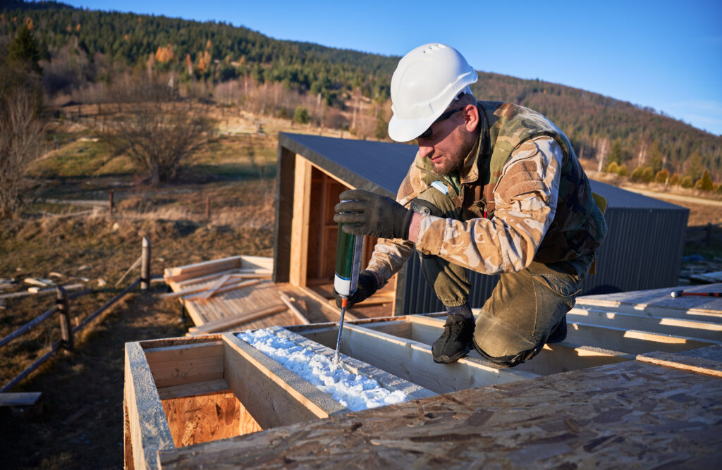 Male builder doing thermal insulation on roof of wooden frame house by polyurethane foam